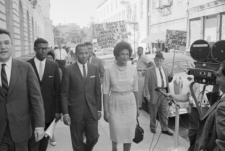 A group of well-dressed people on the sidewalk outside an office building, with picketers in the background.