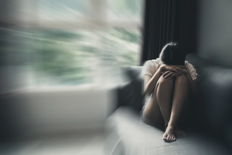 A young woman sits with her head in her knees on a sofa.