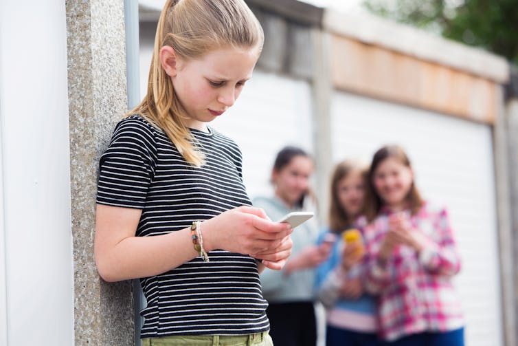 A girl in the foreground looks at her phone while a group of girls behind her laugh.
