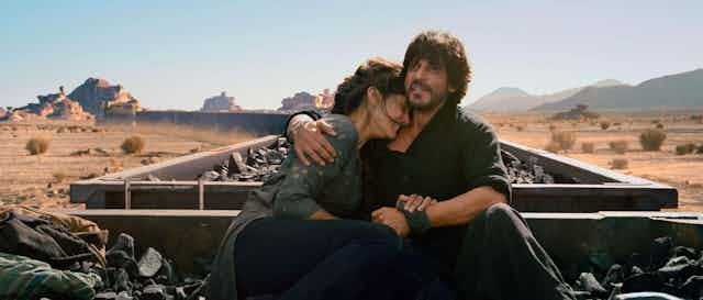 Still from the film Dunki, showing the main characters embracing while sitting on top of open cars on a coal train