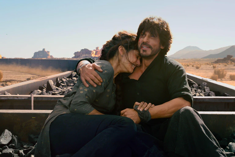 Still from the film Dunki, showing the main characters embracing while sitting on top of open cars on a coal train