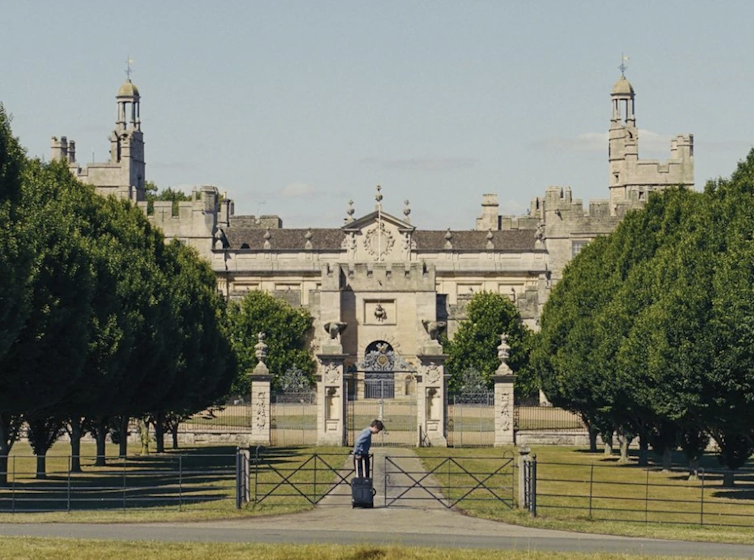 Oliver rolling his suitcase towards a huge stately home.