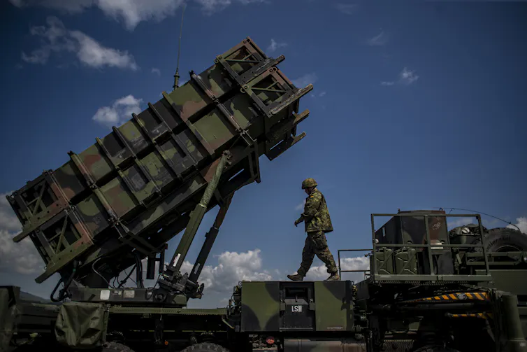 Soldier walks across large military ground vehicle with missile launcher.