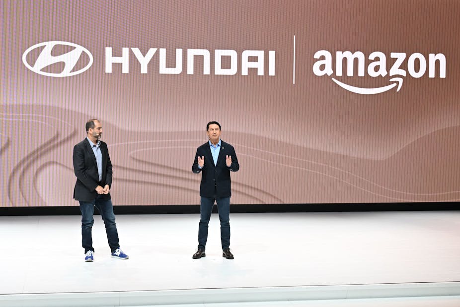 Two executives are seen on a stage. The Hyundai and Amazon logos are visible on a wall behind them.