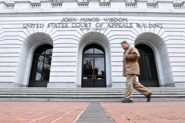 A man dressed in beige walks in front of a large white building with three arched doorways.