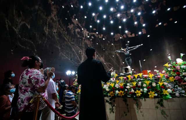 People seen from the back raise their hands in prayer in front of flowers and a crucifix in a large, darkened room.