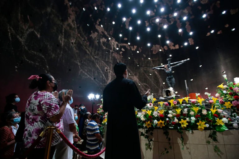 People seen from the back raise their hands in prayer in front of flowers and a crucifix in a large, darkened room.