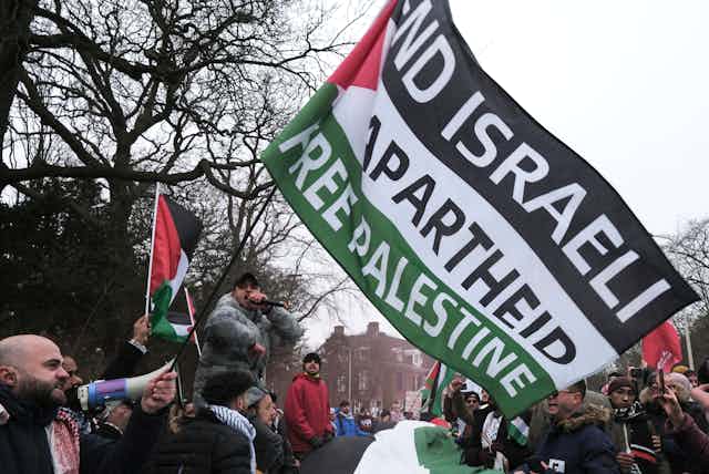 A crowd waves red, white, green and black flags, with one reading End Israeli Apartheid, Free Palestine.