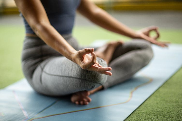 Close-up of person sitting with crossed legs on a yoga mat, hands resting on knees with pointer finger touching thumb