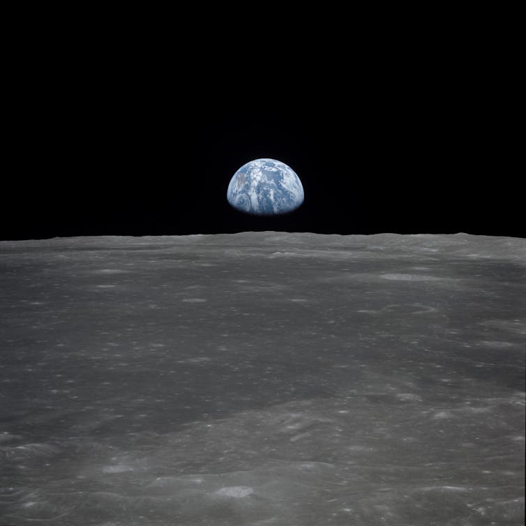 Foreground of a grey surface with a half lit Earth in the distance hanging in a black sky
