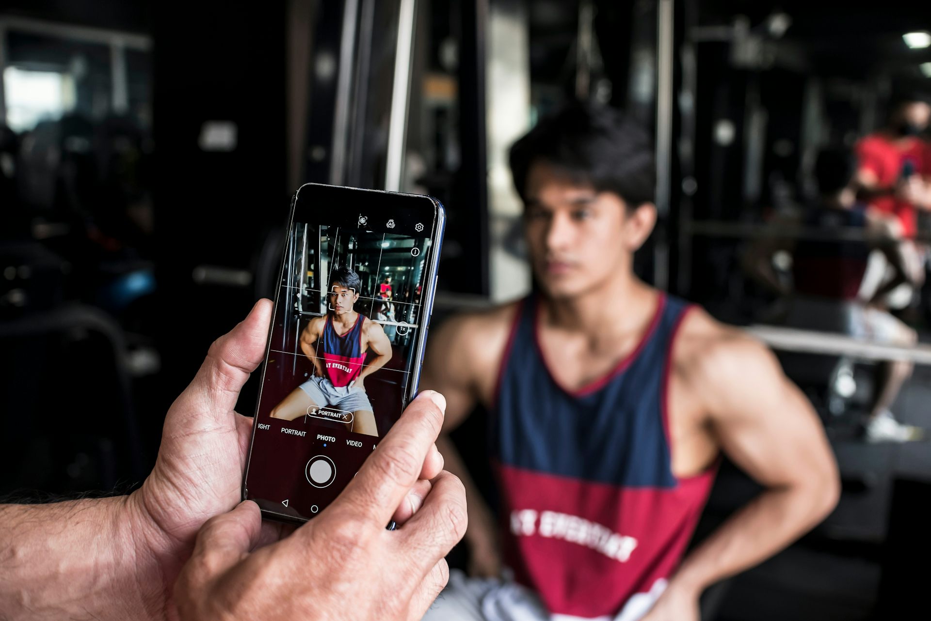 A young man flexes his muscles while someone takes his photo with a smartphone.