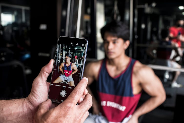 A young man flexes his muscles while someone takes his photo with a smartphone.