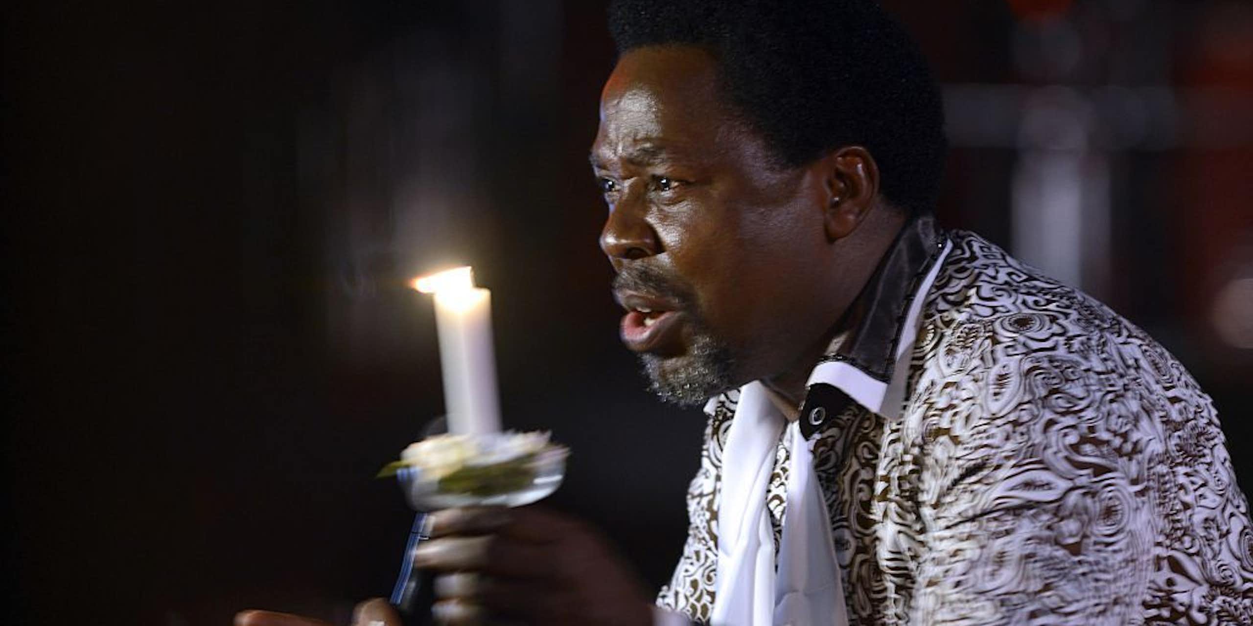 A man holds a candle as he talks, his face earnest. He has a beard and wears an elaborate black and white jacket.
