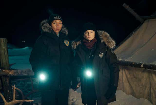 Jodie Foster and Kali Reis dressed as detectives, shining torches in the dark.