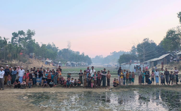 A row of people stood amid waterlogged soil with tin houses in the background.