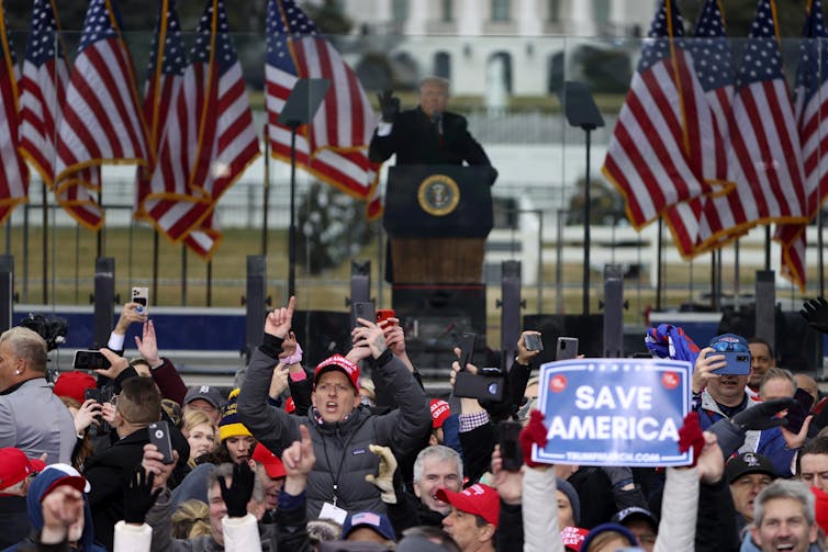 A man in a dark winter coat standing on a stage outside in front of a lot of people, with many American flags behind him.