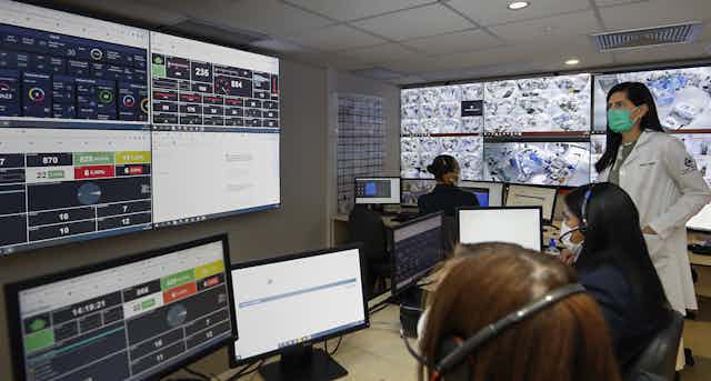 Médica e assistentes trabalham numa sala cheia de computadores e painéis digitais