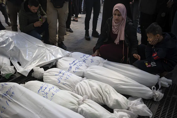 A woman in a pink head scarf stares down at a row of bodies wrapped in white cloth.
