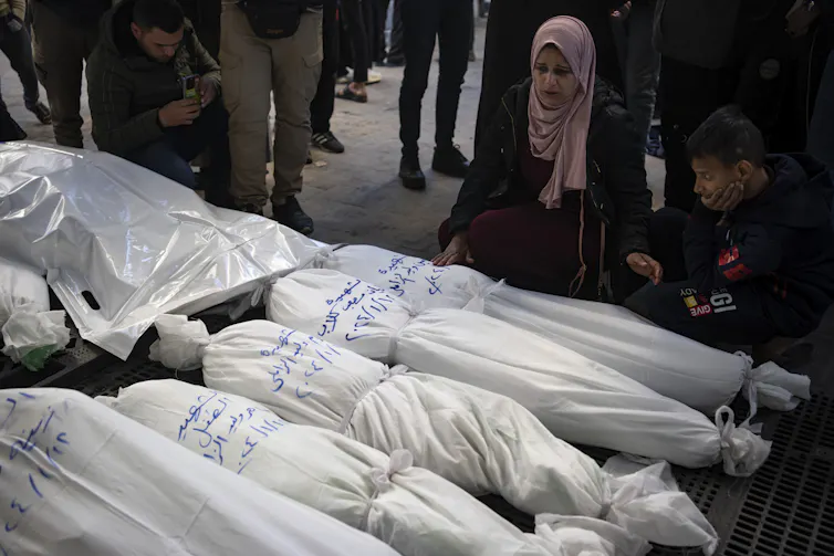 A woman in a pink head scarf stares down at a row of bodies wrapped in white cloth.