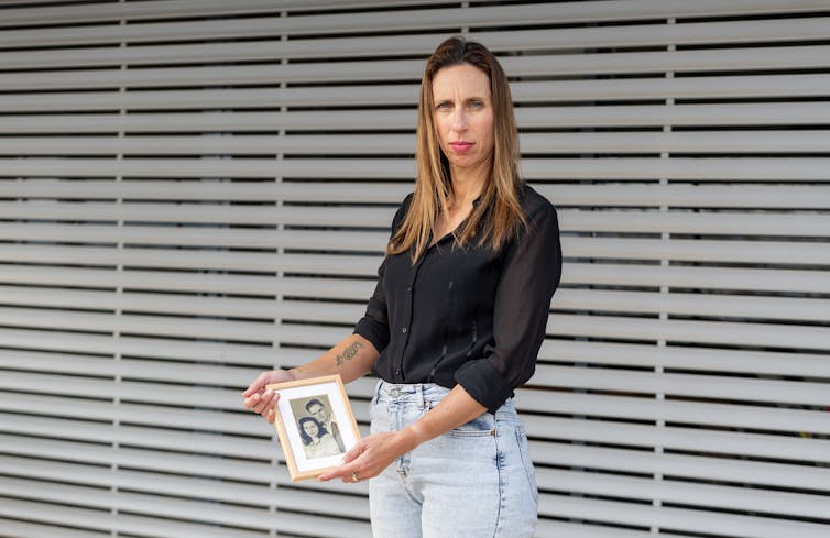 A woman in a black shirt and jeans holds out a framed photograph.