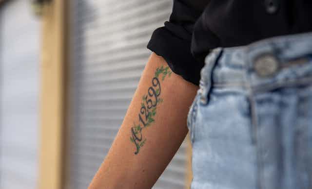 A woman wearing jeans and a black shirt holds out her arm on which is tattooed a number.