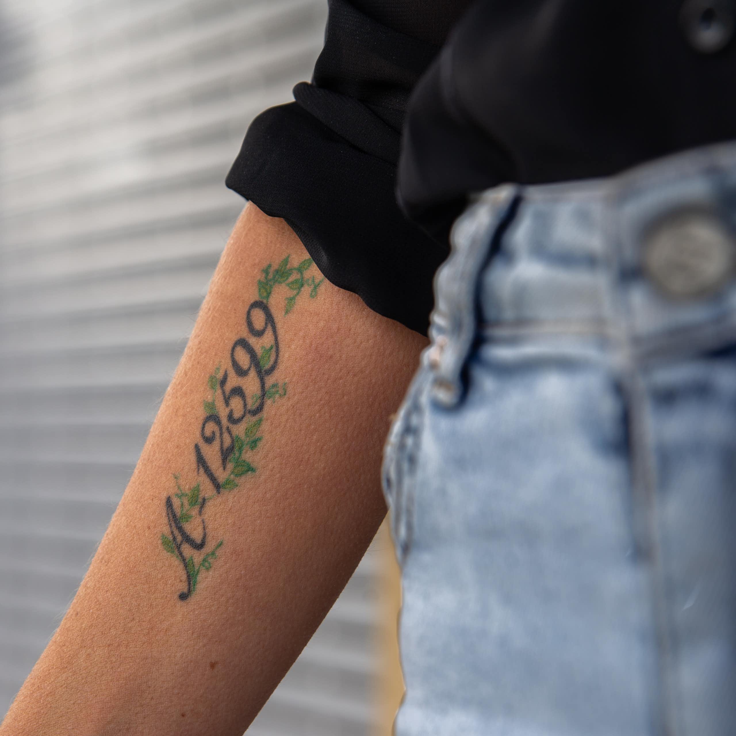 A woman wearing jeans and a black shirt holds out her arm on which is tattooed a number.