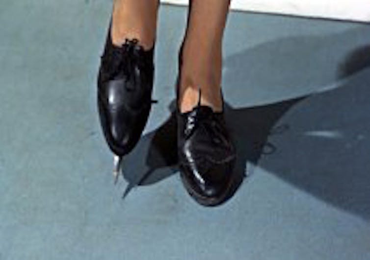 A woman's legs in a pair of shoes with aknife sticking out of the toe of the right foot.