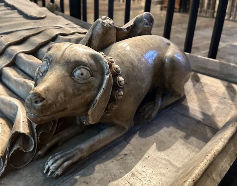 A statue of a dog wearing a collar carved from stone at the feet of a tomb.