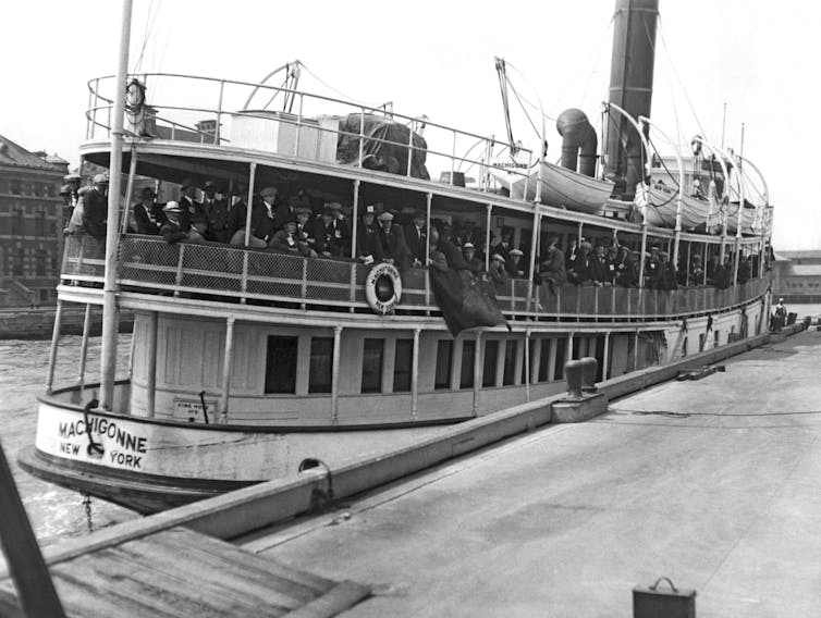 A black and white photo shows a large crowd of people, mostly men wearing hats, crowded on the second story of a small boat, which is coked.