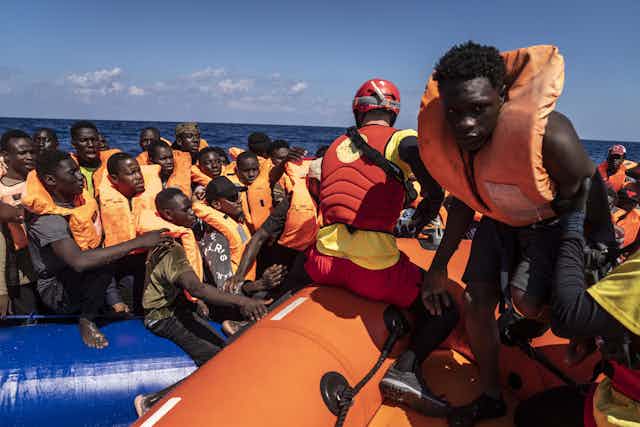 Black men wearing orange life jackets all crowd on a small boat, as one person walks on another adjacent boat and a person wearing a red helmet and vest kneels behind the man walking.