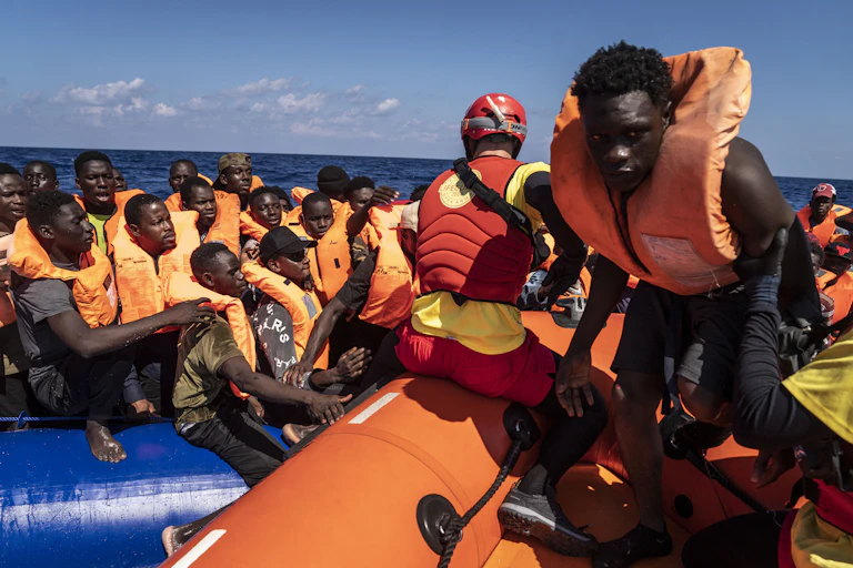 Black men wearing orange life jackets all crowd on a small boat, as one person walks on another adjacent boat and a person wearing a red helmet and vest kneels behind the man walking.