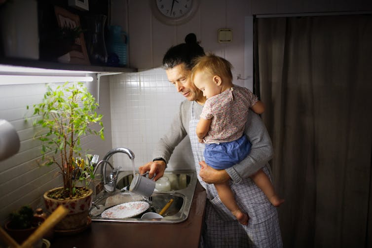 A middle-aged man washing dishes at a kitchen sink while holding a toddler in one arm