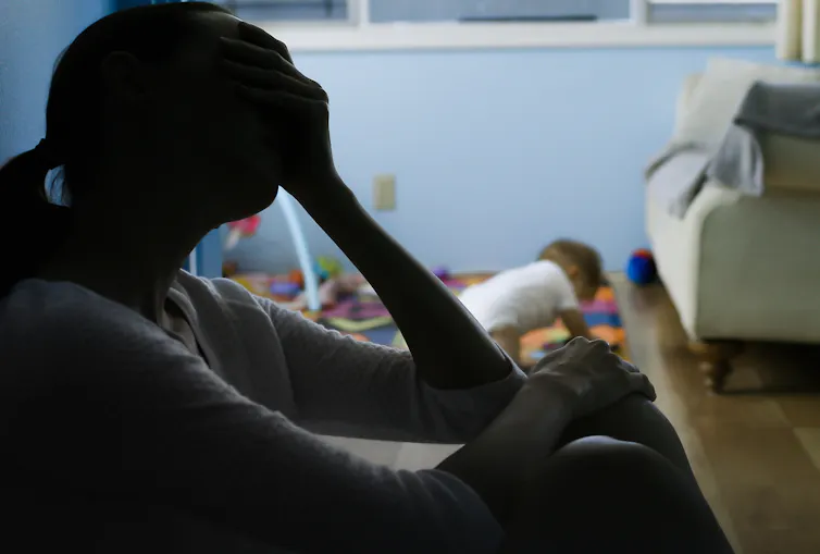 The silhouette of a woman sitting with her head resting against her hand while a toddler plays in the background
