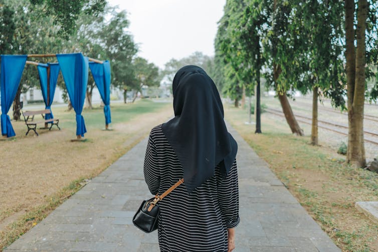 A girl in a black hijab with a handbag walks down a tree-lined path