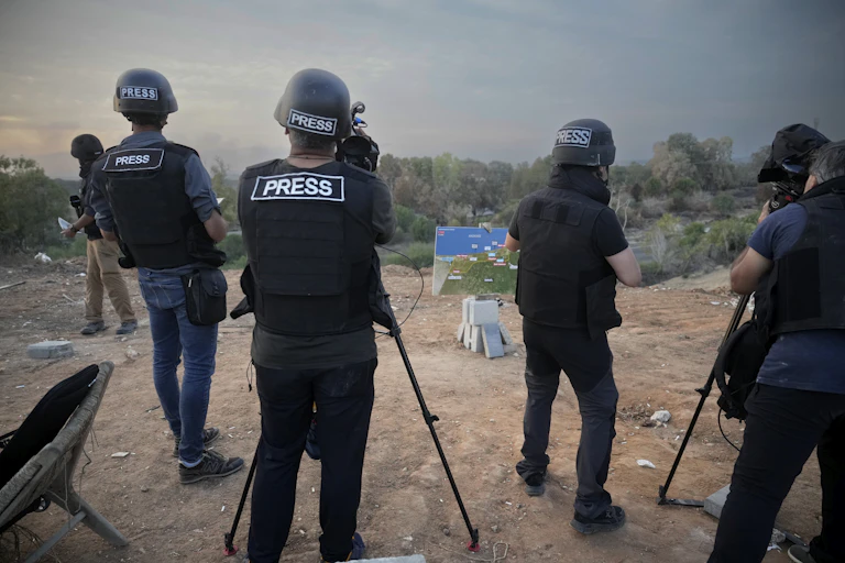 A group of reporters wearing 'press' helmets and vests report in a warzone