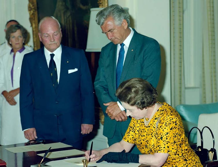 Queen Elizabeth II signing paper at a desk while a man in a suit stands over and watches