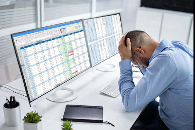 A man sits with his head in his hands in front of two screens containing spreadsheets