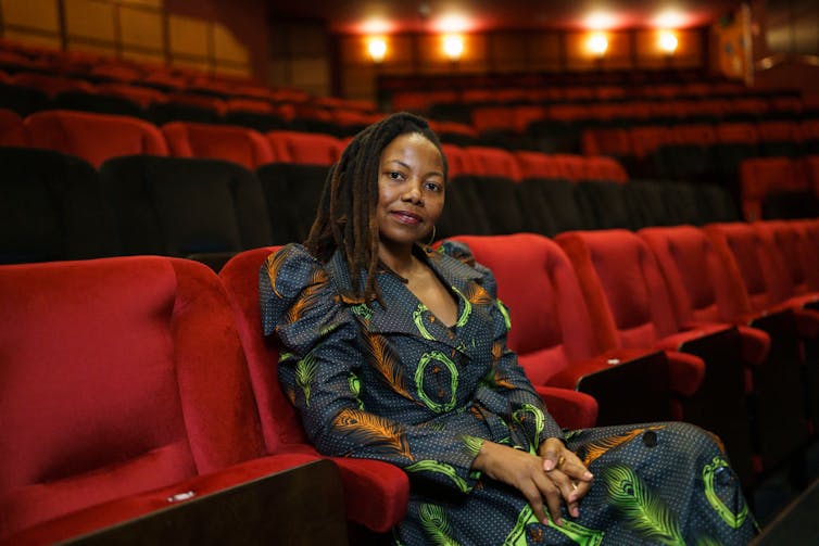 A woman in a dress of African print fabric sits in a theatre with red seats, looking at the camera with a slight smile.