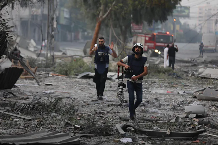 Two journalists wearing jackets saying PRESS walk through devastated area in Gaza.