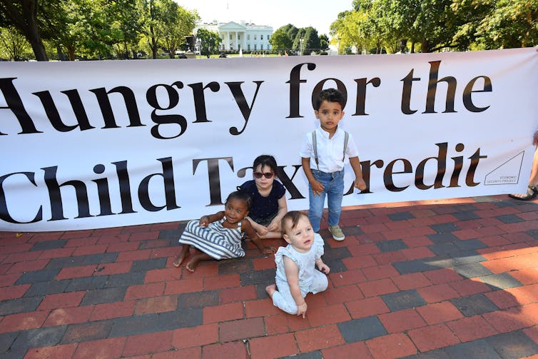 A group of children sit and stand in front of a banner that says 'hungry for the child tax credit.'