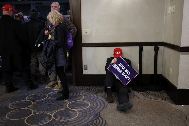 A person sits on the floor of a hotel hallway with a red hat that says USA in white and an upside down campaign banner that says 'Live free or die.' People, some also wearing red hats, walk nearby him.