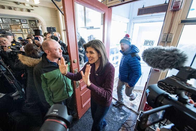 Nikki Haley walks through a doorway, waving with both hands, to a crowd of people. Several video cameras and phones are pointed closely at her.