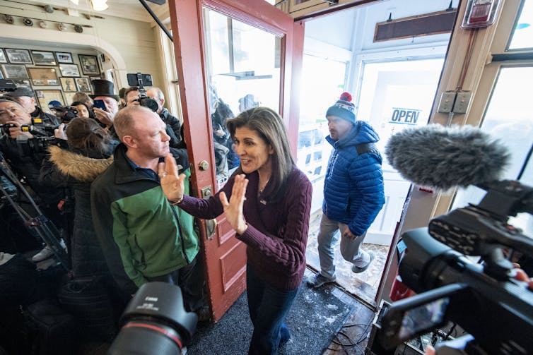 Nikki Haley walks through a doorway, waving with both hands, to a crowd of people. Several video cameras and phones are pointed closely at her.