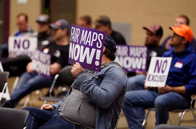 Men wearing baseball caps sit in chairs and hold up signs that read "FAIR MAPS NOW!" and "NO EXCUSES."