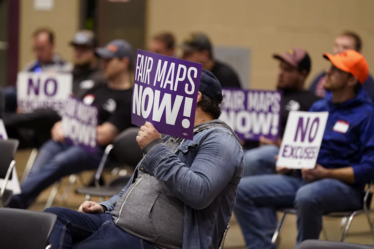 Men wearing baseball caps sit in chairs and hold up signs that read "FAIR MAPS NOW!" and "NO EXCUSES."