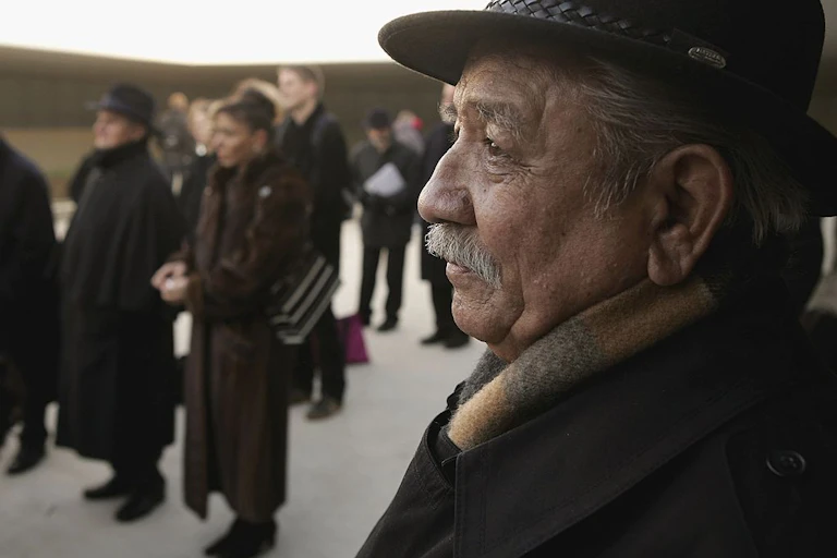 A man in a coat, scarf and hat stands in profile among a thin crowd of people in heavy coats.