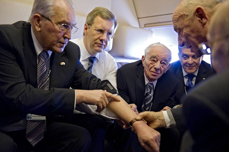 Five older men in suits and ties sit in a semicircle as one pulls up his sleeve.