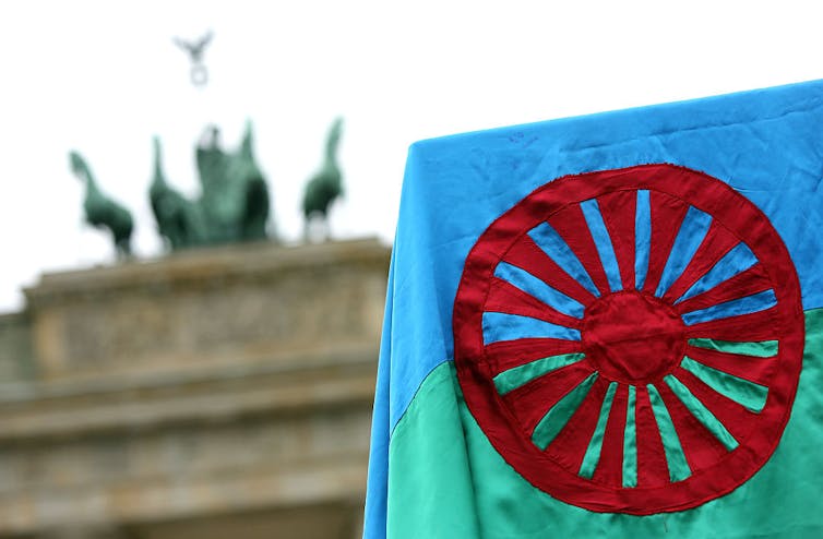 A blue and green flag with a red wheel design waves in front of a large stone monument with a statue on top.