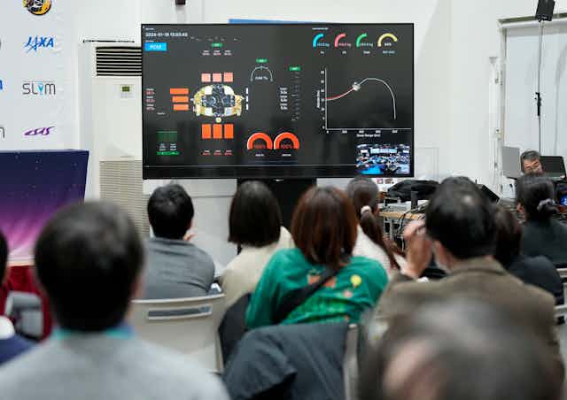 The backs of a crowd of journalists looking up at a screen with several meters and a graph showing the craft's metrics. 