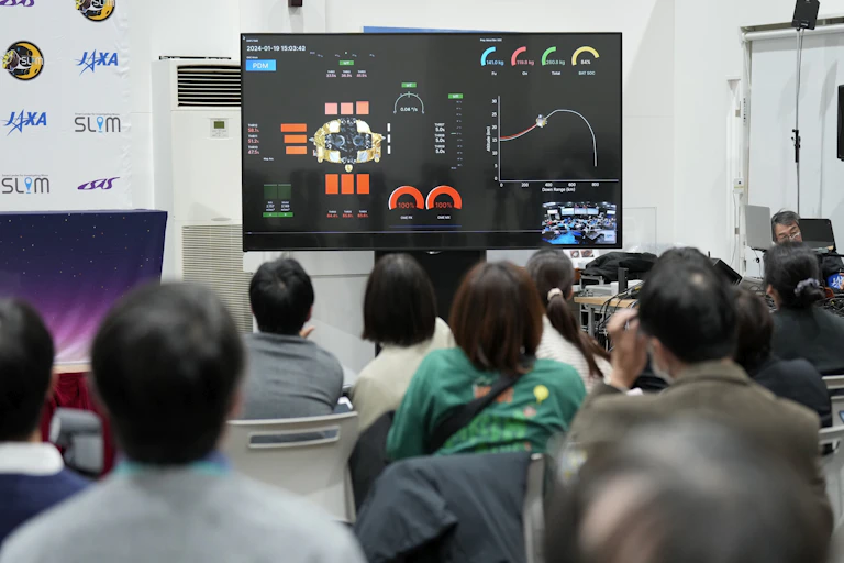 The backs of a crowd of journalists looking up at a screen with several meters and a graph showing the craft's metrics.